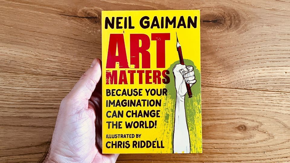 A hand holding a copy of Art Matters by Neil Gaiman, illustrated by Chris Riddell, over a wooden table