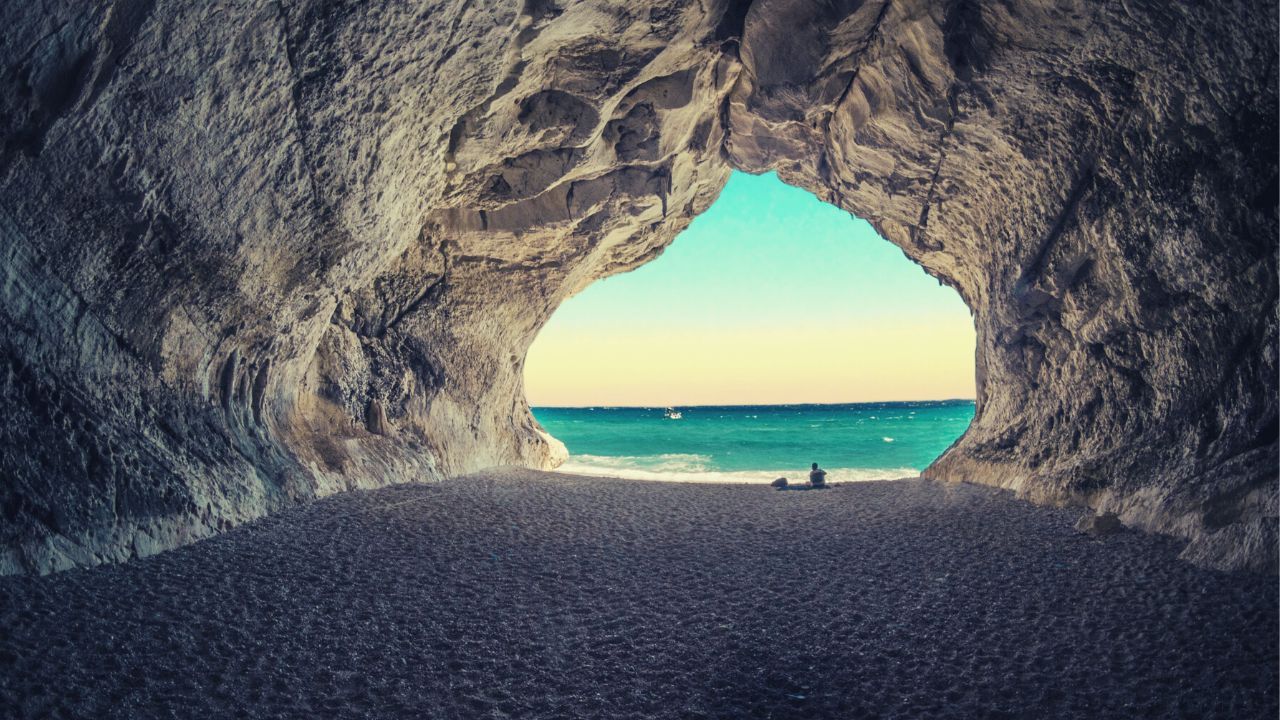View from inside a sea cave towards a turquoise sea, with a person sitting on the sand