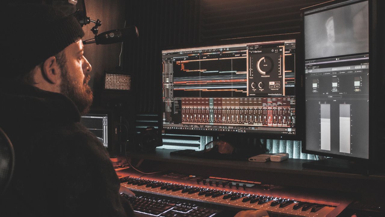 Producer at a studio desk with a MIDI keyboard and monitors showing a DAW and a de-esser plugin